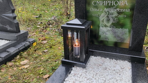 Гранитная лампада зажжённой свечой на фоне вертикального стеклянного памятника